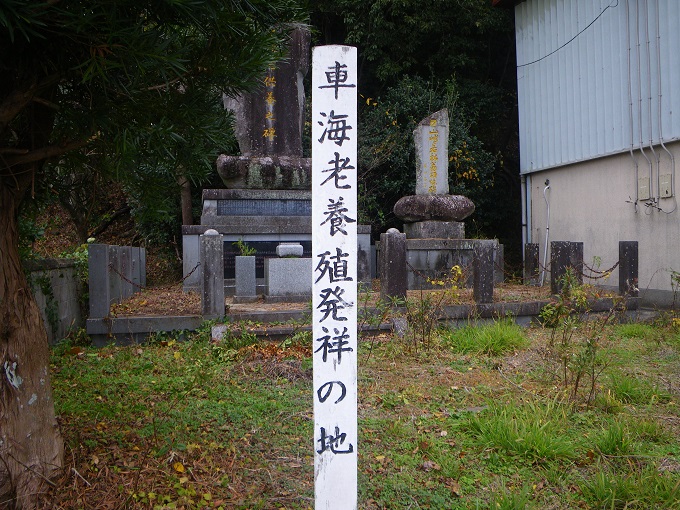 中央手前に「車海老養殖発祥の地」と表示された白い標柱、後方の１メートル程度の台座の上に「車海老供養之碑」と表示されている約２メートルの高さの石碑が祀られている様子を撮影した写真