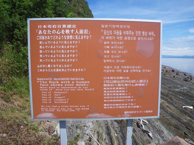 人面岩のある海岸への入口に設置されている、日本語、英語、韓国語、中国語の４カ国語で人面岩の説明が表示されている看板を撮影した写真（説明文　日本奇岩百景認定「あなたの心を映す人面岩」ご対面されてどのような表情に見えますか？悲しんでいるように見えますか？喜んでいるように見えますか？怒っているように見えますか？笑っているように見えますか？叱っているように見えますか？心が少し軽くなりましたか？これからどんな道を歩んでいきますか？）
