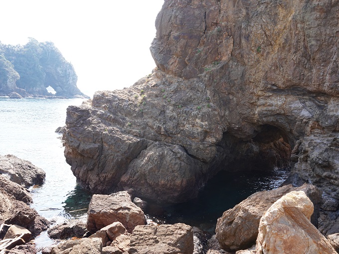 左上奥に妙見岩のゾウの頭の部分が写り込んだ、右下に洞穴がある大きな岩を写した写真で、洞穴の中の海面が光っている。